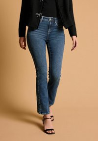 Jean en denim bleu taille haute, coupe slim, ourlet brut et légère décoloration. Associé à des talons noirs à brides. Le fond est de couleur beige clair.