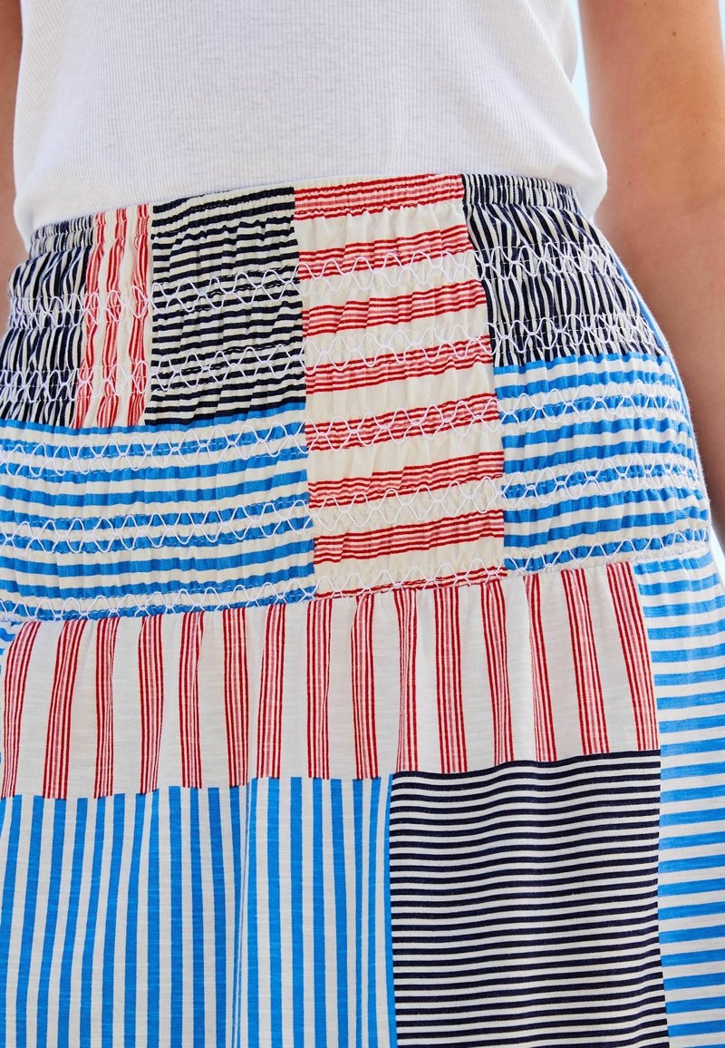 Ceinture d’une jupe en patchwork de rayures rouges, bleues et noires de différentes largeurs, portée avec un haut blanc.