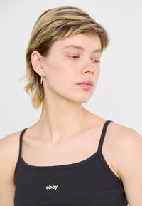 Svart tanktop med tynne stropper, med en liten "obey" logo i gull på forsiden. Glatt tekstur, lett stoff.
