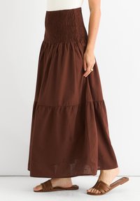 Brauner, gestufter Maxirock mit einem gerafften Bund, aus leichtem Material gefertigt. Das Design zeichnet sich durch eine fließende Silhouette aus und wird mit braunen Riemchensandalen getragen.