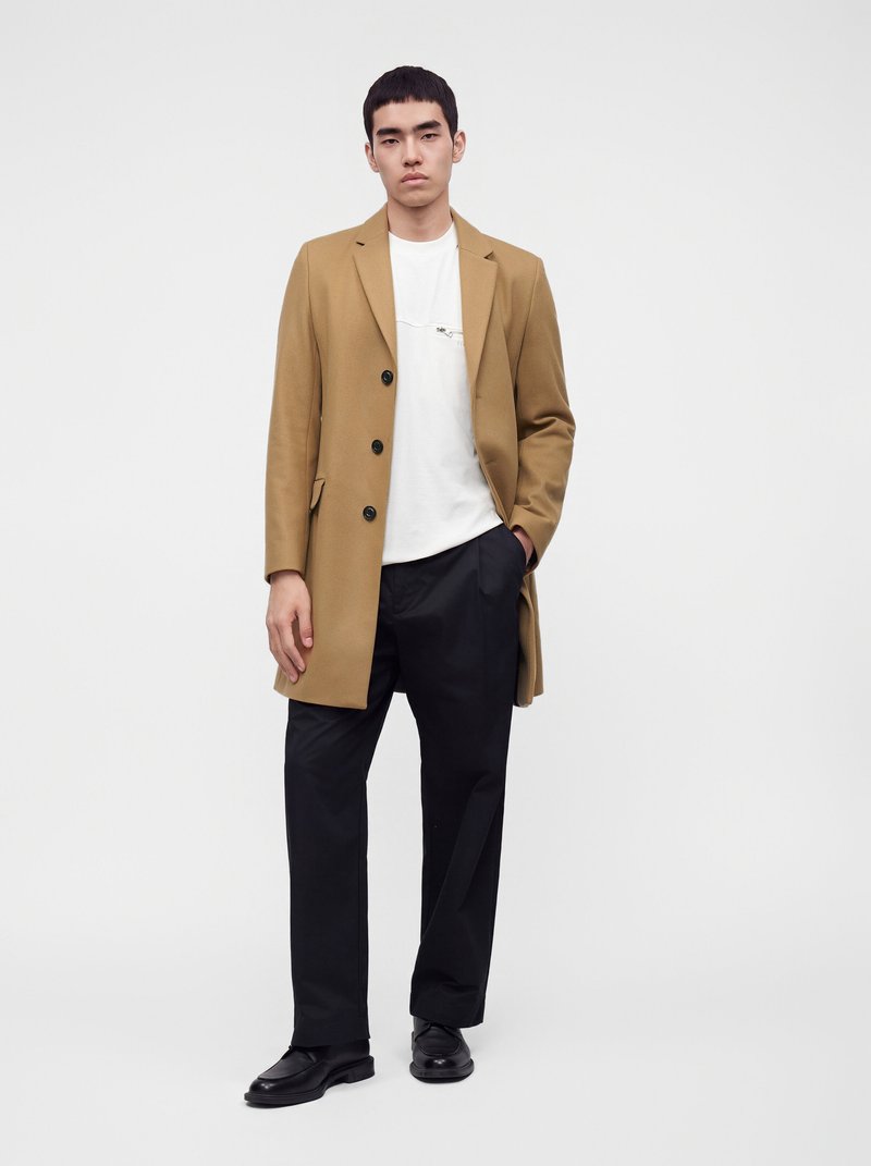 Homme portant un manteau camel boutonné, un t-shirt blanc, un pantalon noir et des chaussures noires, debout devant un fond blanc uni.