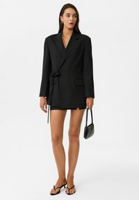 Zwarte blazer jurk met een oversized pasvorm, enkelvoudige knoopsluiting, strik taille detail en twee voorzakken, gecombineerd met een kleine zwarte handtas.