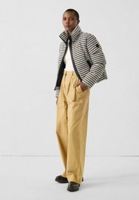Vrouw die staat in een zwart-wit houndstooth pufferjack, witte trui, beige wijde broek en zwarte laarzen tegen een eenvoudige achtergrond.