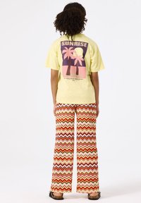 Geel grafisch T-shirt met palmboomontwerp en de tekst "SUNRISE", gecombineerd met wijde zigzag-patroonbroek in rood, roze en geel.