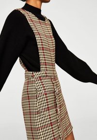 Beige houndstooth-mekko punaisilla yksityiskohdilla, jossa on suuret pyöreät napit ja vartalonmyötäinen muoto. Pitkät mustat hihat näkyvissä.