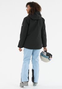 Schwarze wasserdichte Jacke mit Kapuze, kombiniert mit hellblauen Schneehosen. Die Person hält einen Helm mit getöntem Visier und trägt Schneeschuhe.