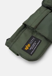 Borsetta verde in nylon con chiusura a zip, dotata di patch con logo, patta pieghevole e tasca aggiuntiva, adatta per vari accessori.