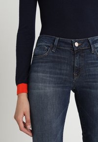 Granatowe dżinsy o dopasowanym kroju; cechy to zapięcie na guzik, kieszenie z przodu oraz lekkie przetarcia na udach. Czerwony akcent w nadgarstku.