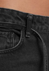 Lähikuva mustista denim-farkuista, joissa on vyölenkit, pieni etutasku ja näkyvä yläommellus kankaassa.