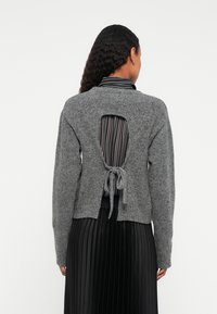 Donna con lunghi capelli ricci indossa un maglione grigio a maglia con schiena scoperta legato in vita sopra una camicia a righe e una gonna nera plissettata.