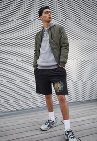 Olivfarbener Bomberjacke über einem grauen Hoodie, schwarze Shorts mit grafischem Druck, weiße Socken und mehrfarbige Sportschuhe auf Holzfußboden.