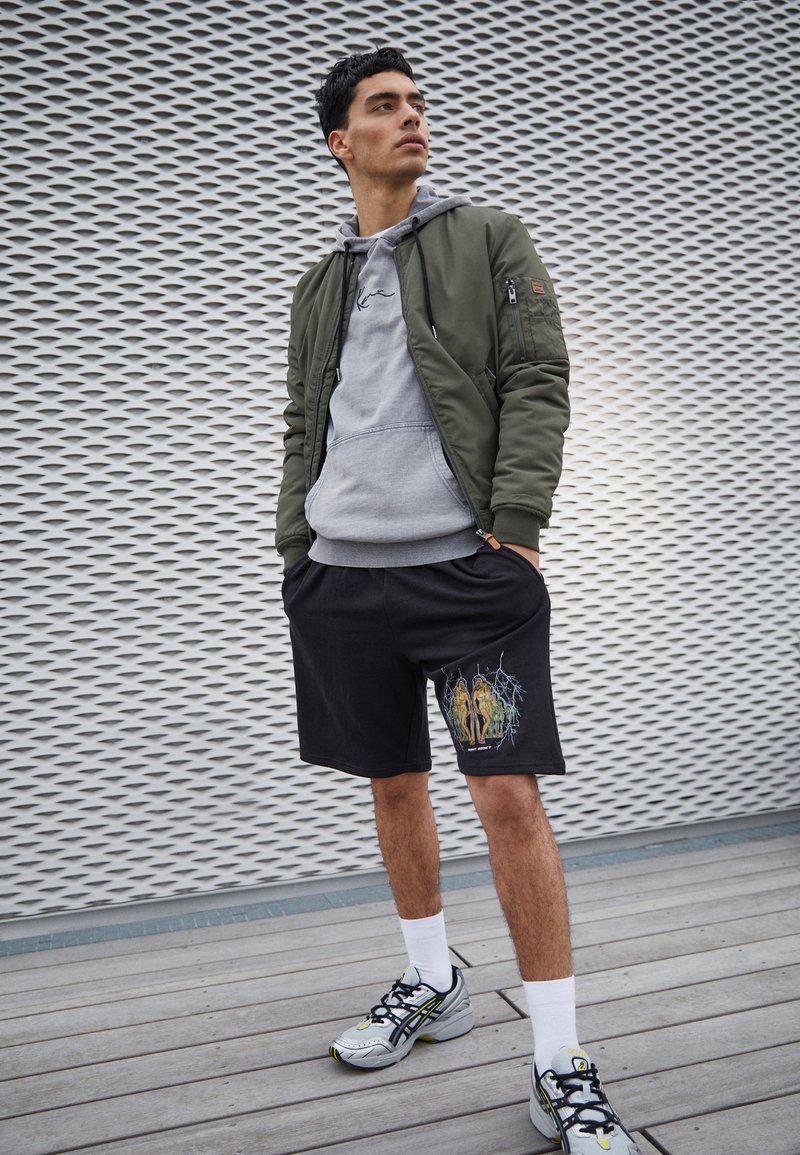 Olivfarbener Bomberjacke über einem grauen Hoodie, schwarze Shorts mit grafischem Druck, weiße Socken und mehrfarbige Sportschuhe auf Holzfußboden.