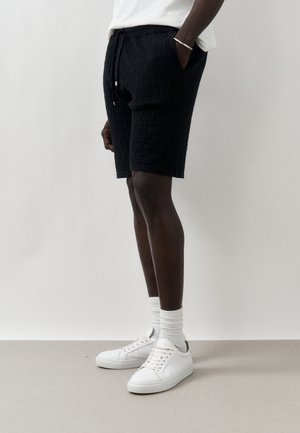 Mand stående iført teksturerede sorte shorts, hvide crew-sokker, hvide sneakers og en hvid skjorte med den ene hånd i lommen.