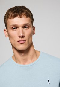 Lichtblauwe sweater met ronde hals, gemaakt van gebreide stof, met een klein marineblauw logo aan de linkerkant, een gladde textuur en een eenvoudig ontwerp.