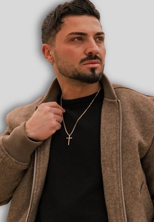 Braune Zip-Jacke mit gerippten Bündchen über einem schwarzen Pullover. Goldene Kreuzkette, die mit einer Hand gehalten wird. Nahaufnahme-Porträt mit neutralem Hintergrund.