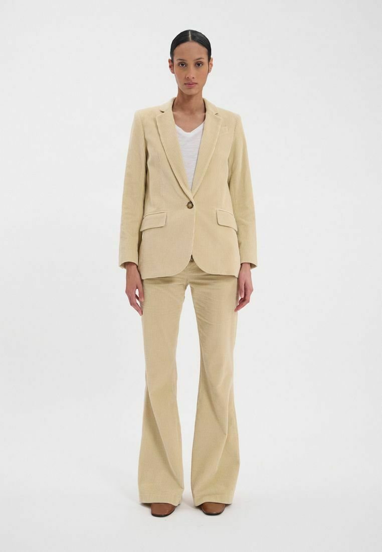 Costume en velours côtelé beige avec blazer ajusté et pantalons évasés. Le blazer possède un bouton unique et des poches avant ; texture douce dans l'ensemble.