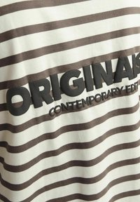 Röstrikkiga puuvillane särk, millel on beežid ja pruunid horisontaalsed triibud. Suur must tekst "ORIGINALS" allpool sõnadega "CONTEMPORARY EDIT".