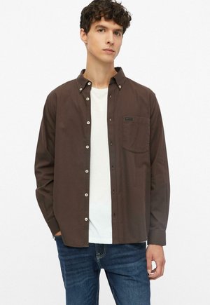 Junger Mann, der ein dunkelbraunes Hemd über einem weißen T-Shirt und blauen Jeans trägt, steht mit einer Hand in der Tasche vor weißem Hintergrund.