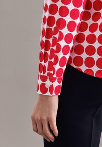 Koszula z długim rękawem o białej podstawie z dużymi czerwonymi kropkami. Posiada guzikowe mankiety oraz dopasowaną konstrukcję o gładkiej teksturze.