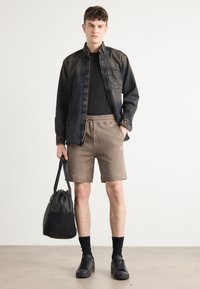 Chemise en denim noir, t-shirt noir, shorts khaki, baskets noires, chaussettes noires. Tenant un sac noir. Tenue simple et décontractée sur une silhouette masculine.