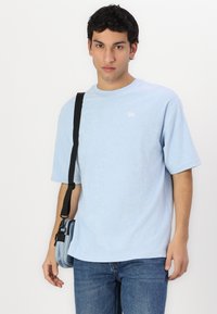 Gaiši zils īsām piedurknēm T-krekls, izgatavots no mīksta auduma, ar apaļu kakla izgriezumu un mazu logo. Valkāts ar crossbody somu un džinsiem.