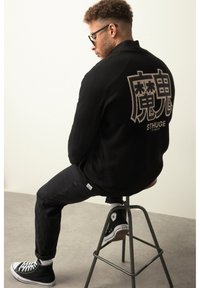 Sort fleecejakke med stort grafisk motiv på ryggen, parret med mørke bukser og sorte høj-top sneakers, siddende på en skammel.