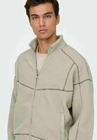 Giacca in pile beige con colletto alto, zip frontale e dettagli delle cuciture a contrasto. Presenta tasche laterali e materiale morbido e testurizzato.