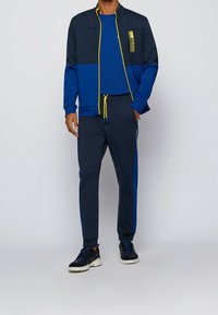 Sportovní komplet v námořnické modři a modré barvě. Bunda má vysoký límec, zapínání na zip, žluté akcenty a boční kapsy. Kalhoty jsou zúžené s modrými pruhy po stranách.