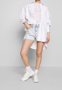 Witte katoenen shirt over een lichtbeige t-shirt, gecombineerd met lichtblauwe denim shorts en chunky witte sneakers. Eenvoudig ontwerp en een relaxte pasvorm.