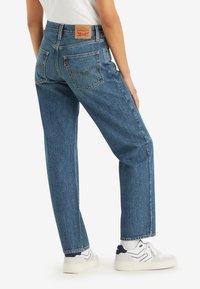 Blue denim jeans with a loose fit, featuring a high waist, two back pockets, and a brown leather logo patch at the waistband.