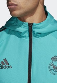 Turquoise sportjack met capuchon, voorzien van een zwarte rits, zwart Adidas-logo en Real Madrid-embleem op de borst. Gestructureerde stof.