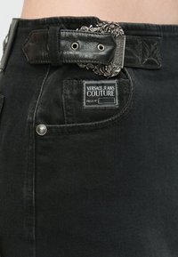 Fekete farmernadrág textúrált fekete bőrpánttal, részletes ezüst csattal és márkázott címkével a hátsó zsebében.