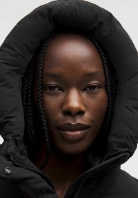 Sort puffer-jakke med en overdimensioneret hætte, der har lynlås-lukning og struktureret stof. Glat hud og fletninger er synlige.