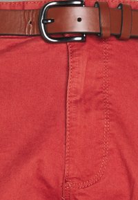 Pantalon en tissu rouge avec une texture lisse, arborant une ceinture en cuir marron avec une boucle en argent. Coutures visibles le long des coutures.