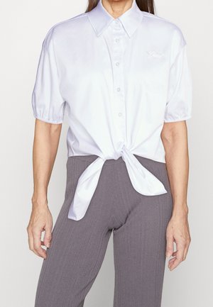 Kobieta ubrana w białą koszulę na krótki rękaw zapinaną na guziki, związaną w talii, w szare prążkowane spodnie, stojąca na tle jasnego, jednolitego tła.