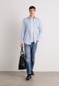 Camisa de botones a rayas azul claro, camiseta blanca debajo, jeans azules, zapatos negros y una bolsa de tela negra, de pie frente a un fondo liso.