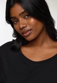 Czarny t-shirt z szerokim dekoltem, noszony przez osobę z długimi, ciemnymi włosami. Odcień skóry średni, gładka tekstura.