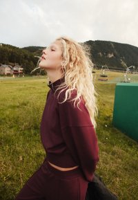 Bordová krátka mikina s dlhými rukávmi, doplnená o zladené tepláky. Vlnité svetlé vlasy; zelené pozadie s trávou a vzdialenými budovami.