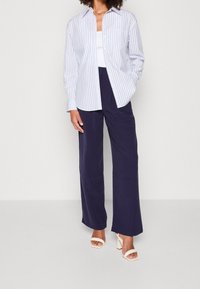 Wit-blauw gestreept overhemd met knopen en lange mouwen, gecombineerd met losse donkerblauwe broek en lichte sandalen met blokhak.