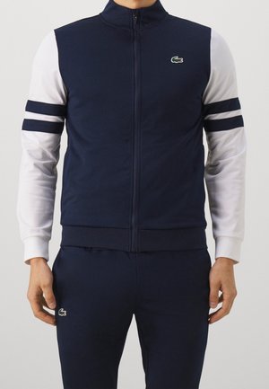 Marineblauer Reißverschluss-Pullover mit weißen Ärmeln, jeweils zwei marineblaue Streifen an den Armen und ein kleines grünes Krokodil-Logo auf der Brust. Weiche Textur.