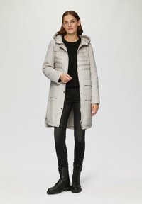 Lichtgrijze geïsoleerde jas met een capuchon, knopen aan de voorkant en opgestikte zakken. Draagt over een zwart gestructureerd sweater en zwarte skinny jeans, gecombineerd met zwarte laarzen.