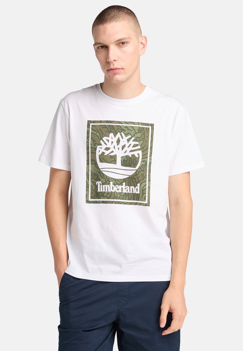 Camiseta de algodón blanca con un logotipo de árbol en camuflaje verde en un diseño rectangular y el texto "Timberland" debajo. Mangas cortas y cuello redondo.