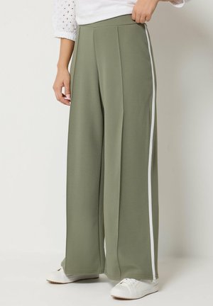 Vrouw draagt olijfgroene wijde broek met witte zijstrepen, witte sneakers en een wit topje met oogjesmouwen, staand tegen een effen achtergrond.