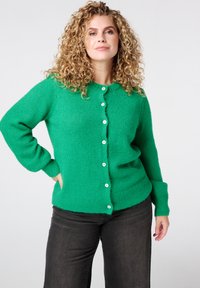 Groene gebreide cardigan met lange mouwen, ronde halslijn en witte knopen. Zachte textuur met een getailleerde tailleband en twee zijzakken.