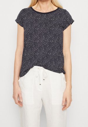 Femme portant un haut noir à manches courtes avec des pois blancs et un pantalon blanc avec cordon de serrage, visible des épaules aux hanches.