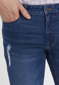 Blå denimjeans med en tettsittende design, med en sidelomme og en slitt detalj nær låret. Kontrastsøm er synlig.
