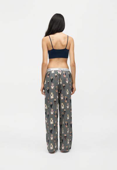 Femme aux longs cheveux foncés, portant un crop top bleu marine et un pantalon de pyjama rayé avec des imprimés d'ours en peluche, debout de dos sur un fond uni.