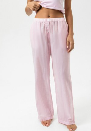 Vrouw draagt lichtroze loszittende pyjamabroek met strepen en knopen aan de tailleband, staand op blote voeten tegen een witte achtergrond.