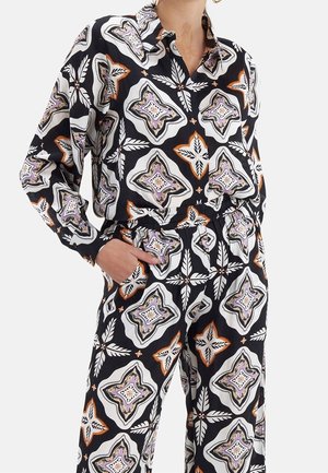 Frau trägt ein passendes schwarzes Outfit mit weißen, orangefarbenen und violetten geometrischen Blumenmustern, Hemd in Gummibundhose gesteckt.