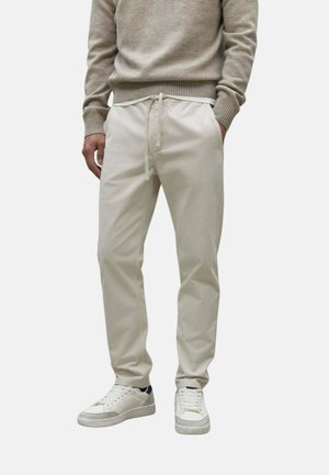 Hombre con pantalones beige con cordón, suéter gris claro y zapatillas blancas, de pie con las manos en los bolsillos.